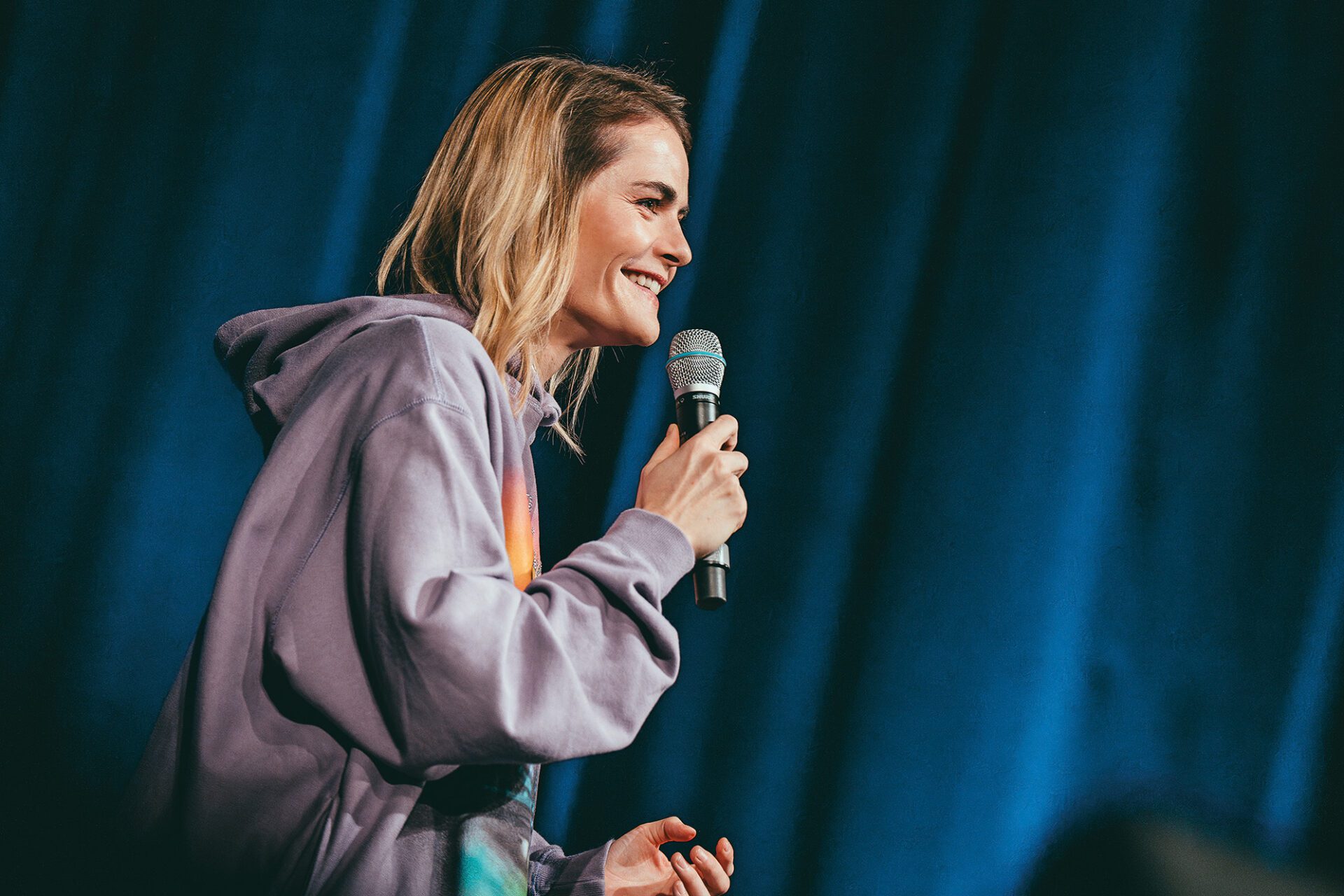 hazel-brugger-c-michael-kramer-web-2 Hazel Brugger mit Mikrofon, lächelnd. Die Komikerin trägt einen Kapuzenpullover.