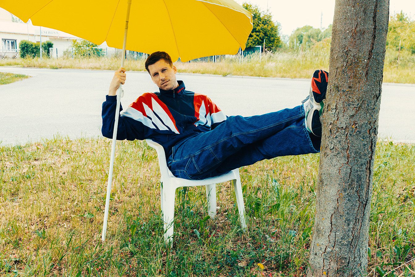 björnvonmorgenstern_KVF_MOTIV1_web Björn von Morgenstern KVF Motiv: Mann mit gelbem Schirm, Tracksuit und Jeans entspannt auf einem Stuhl an einem Baum.