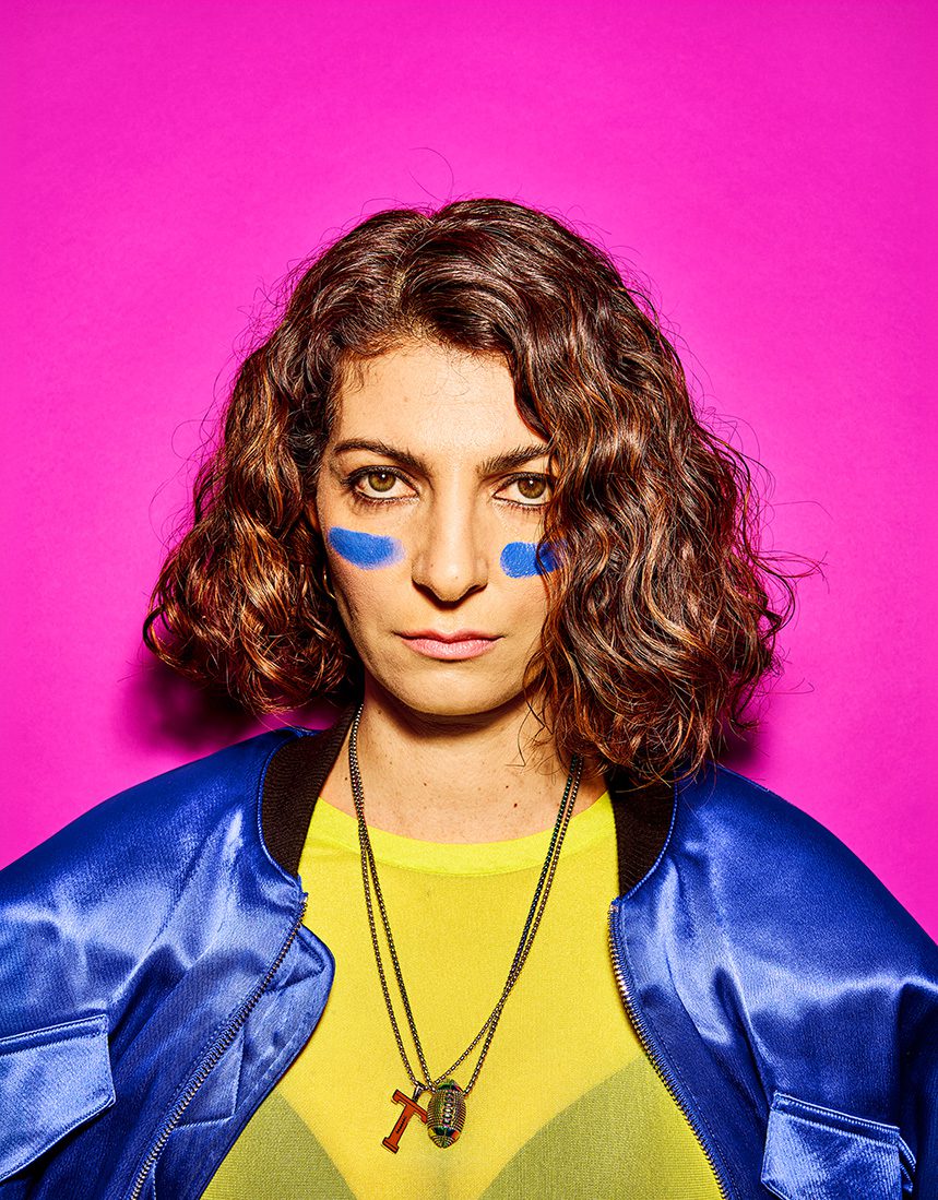 Filiz Tasdan portrait: Woman with blue paint on cheeks against a bright pink background, wearing a blue jacket and necklace.