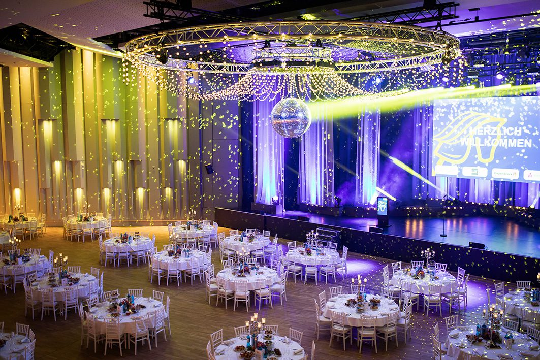 Veranstaltungsbeispiel: Saal mit runden Tischen, Bühne, Discokugel und festlicher Beleuchtung für eine Veranstaltung.