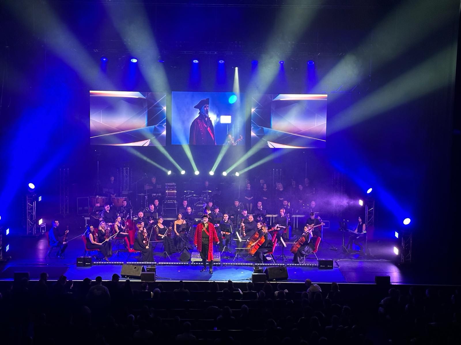 Hans Zimmer 1 (c) Star Entertainment 2024 Ein Dirigent in rotem Mantel leitet ein Orchester auf einer Bühne mit blauer Beleuchtung und grossen Videoleinwänden