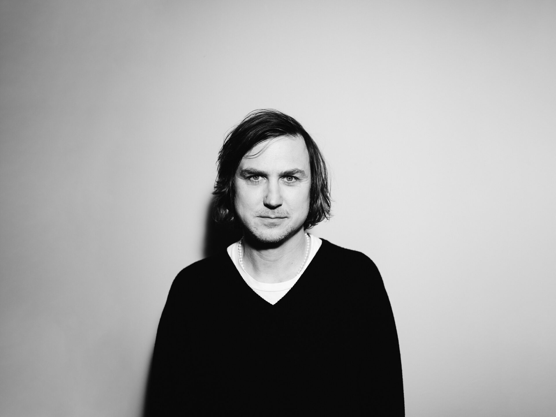 Lars Eidinger by Ingo Pertramer_web Schwarz-Weiß-Porträt des Schauspielers Lars Eidinger mit mittellangem Haar und Perlenkette vor hellem Hintergrund