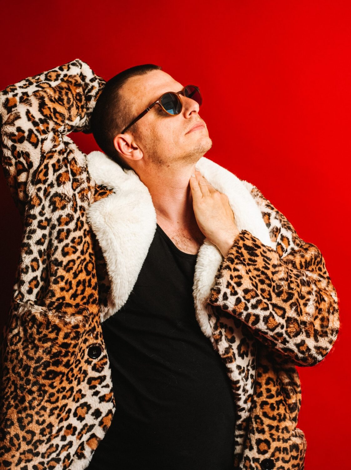 2027_10_04 OsHalle Moritz Neumeier – Pur – Daniel Dittus_web Moritz Neumeier in einem Leopardenmantel mit weißem Fellkragen und Sonnenbrille vor einem leuchtend roten Hintergrund