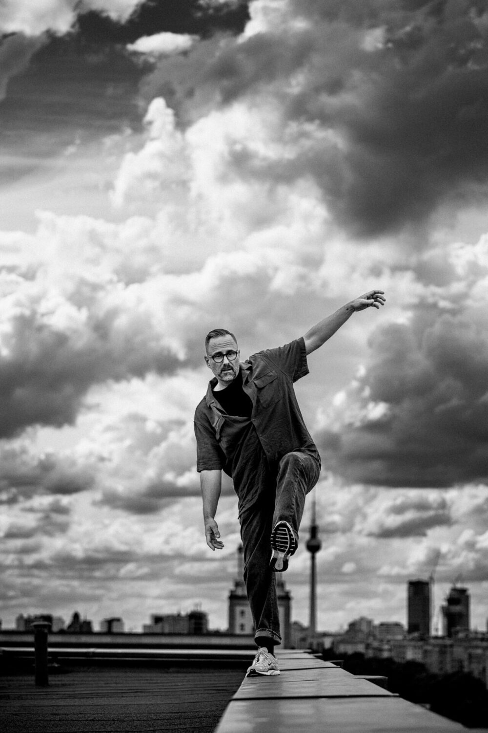 Kurt Krömer_Pressebild Kurt Krömer balanciert in Schwarz-Weiß auf einem Dach mit dramatischem Wolkenhimmel